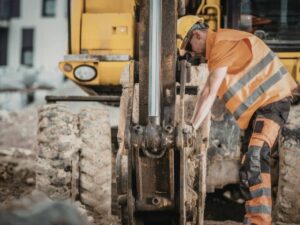 Construction worker in an orange safety vest and hard hat inspecting a hydraulic excavator on a muddy site.